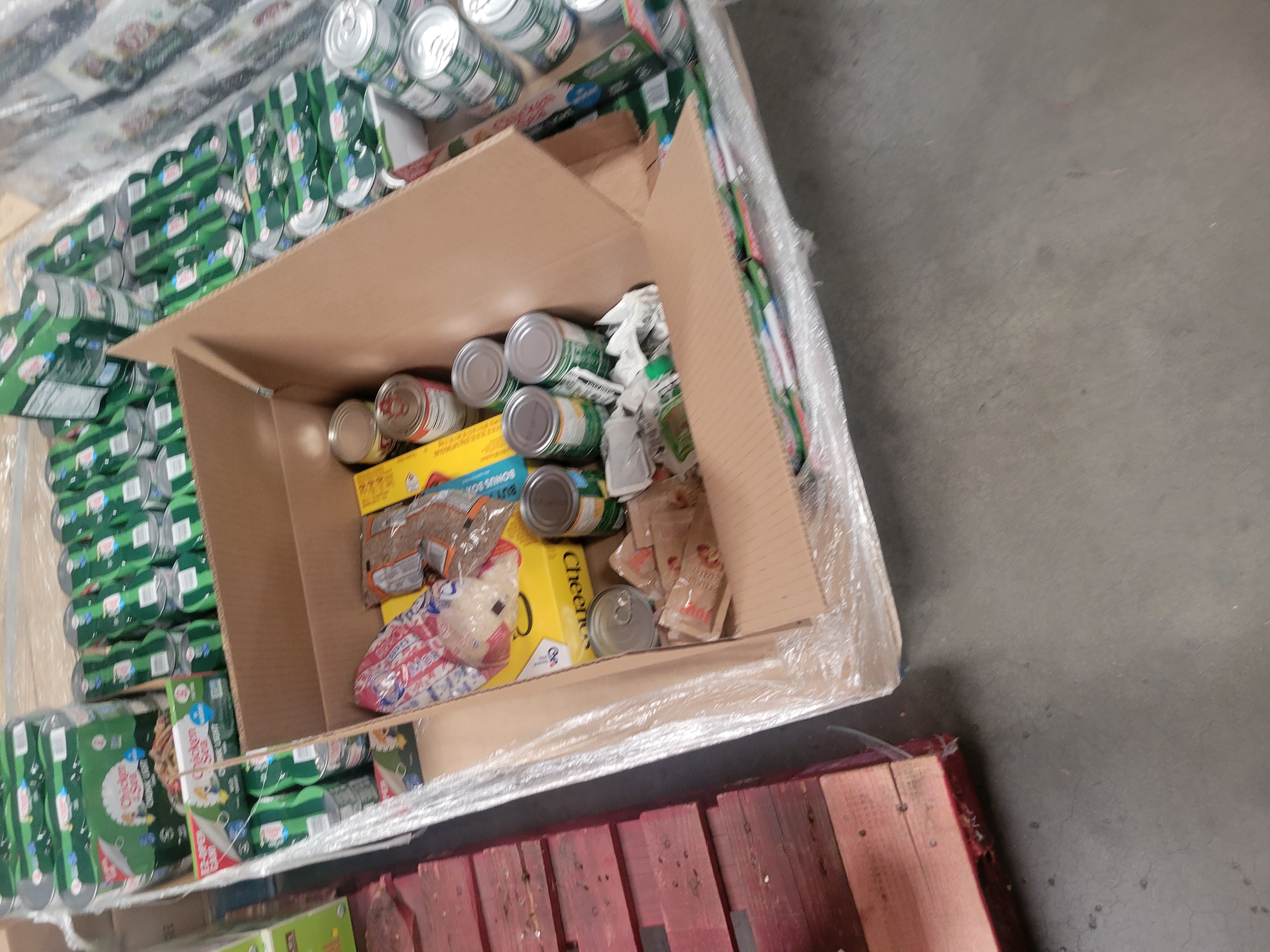 Cardboard box filled with dry goods surrounded by boxes of canned tuna on a pallet covered by plastic wrap.